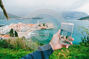 Man& x27;s hand holds a smartphone with a photo of architecture. Left - the old town of Budva, Montenegro