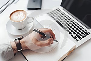 A man\'s hand making a note in a notepad at a white table, next to a MacBook and a cup of coffee. photo
