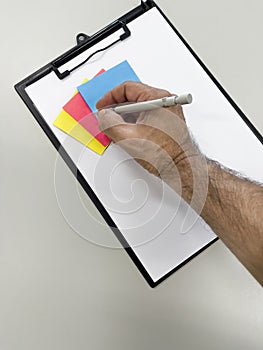 Man`s hand with a metal pen, taking notes in a clipboard papaer on top of yellow, pink and blue memo paper, selective focus
