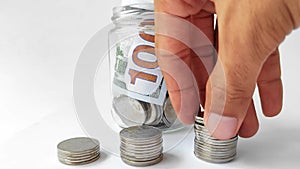 A man\'s hand is placing coins in layers with a background of 100 dollar bills in a glass jar.