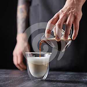 A man`s hand pours coffee into a cup of milk on a black wooden table. Cappuccino preparation.