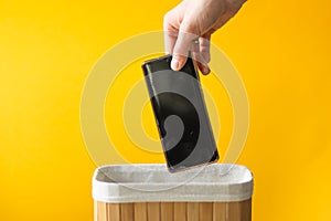 Man's hand putting smartphone into wooden basket on a yellow background. Dependance and digital detox concept.