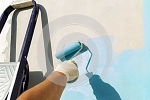 A man`s hand with a roller paints the wall with blue paint. Nearby is a staircase