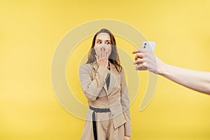 Man`s hand shows a smartphone to a woman, she covers her mouth in shock and looks at the screen. Isolated on yellow background