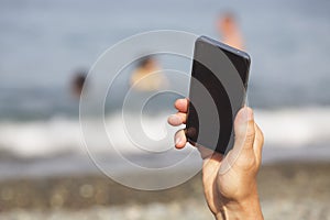 Man's hand with smartphone against the sea and pebble beach. Close-up