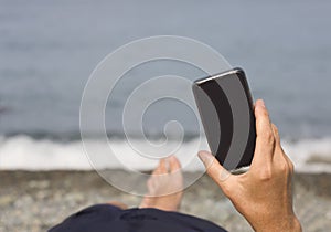 Man& x27;s hand with smartphone against the sea and pebble beach. Close-up