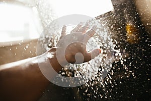 A man`s hand in a spray of water in the sunlight against a dark background. Water as a symbol of purity and life