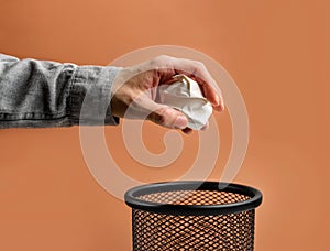 Man`s hand throws crumpled paper into a basket on a beige background with copy space
