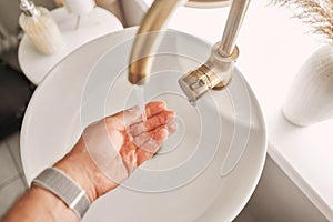 A man& x27;s hand touches the water in a beautiful sink with a golden faucet next to an oval mirror and a shelf with hand