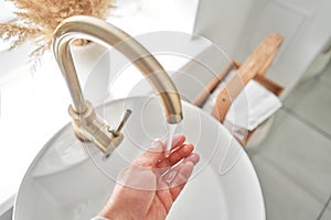 A man& x27;s hand touches the water in a beautiful sink with a golden faucet next to an oval mirror and a shelf with hand