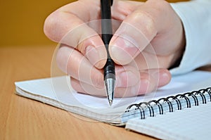 A man`s hand in a white shirt with a black pen in a notebook with a spiral on a wooden table side view closeup