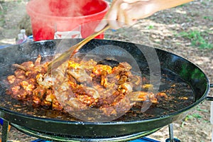 Man`s hand with wooden turner mixing fried chicken meat with tomato sauce in large flat pan. Preparing paella or jambalaya