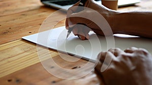 A man`s hand writing on a white paper on a table with a cup of coffee and a computer notebook nearby