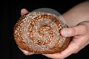 Man`s hands hold tasty fresh loaf of dark bread with sesame seeds on black background