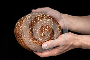 Man`s hands hold tasty fresh loaf of dark bread with sesame seeds on black background