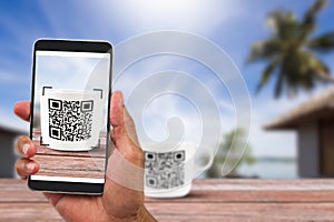 Man`s hands holding smartphone scanning QR code on coffee cup beside the beach