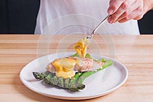 Man`s hands pouring hollandaise sauce on top of delicious baked salmon with steamed green asparagus