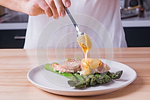 Man`s hands pouring hollandaise sauce on top of delicious baked salmon with steamed green asparagus