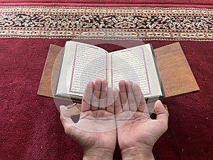 A man\'s hands praying against the background of the Koran and a red carpet