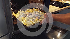 Man's hands put tray with chicken legs and potato slices in the oven, close up, camera tracking