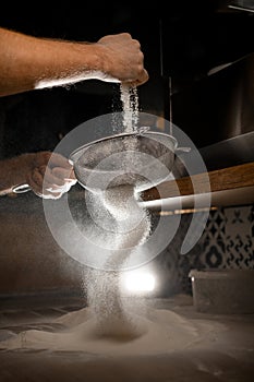 Man's hands sift flour through a metal sieve in a dark kitchen