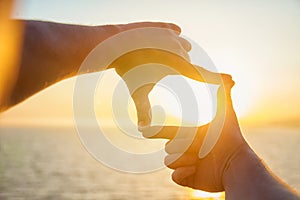 The man`s hands stretch toward the sun on the sea. A man looks at the sunset through the folded fingers.