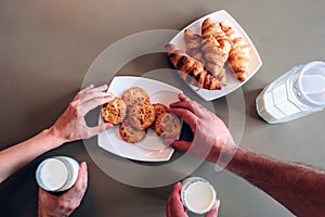 Man`s and woman`s hands are reaching cookies on plate. Also they hold glasses of milk. Isolated on brown background.