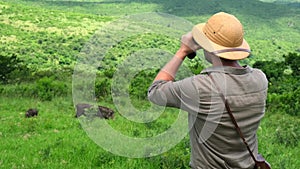 a man in safari clothes looks at the rhinos through binoculars in the savannah