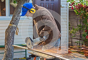A man in the safety glasses and yellow ear protectors and a brown jacket cuts metal using the big white angle grinder