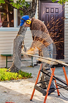 A man in the safety glasses and yellow ear protectors and a brown jacket cuts metal using the big white angle grinder