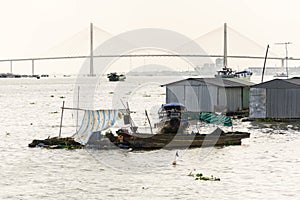 Man sails on ratty boat with fish farm raft houses floating on Mekong river with Rach Mieu Bridge in background