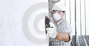 Man sand the wall with sandpaper, professional construction worker with mask, safety hard hat, gloves and protective glasses.