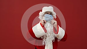 Man in a Santa costume wears a protective mask on his face to protect himself from the coronavirus on a red background. Santa