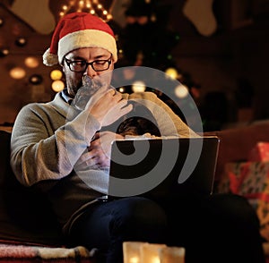 A man in Santa`s New Year hat holds a cat and working with laptop.