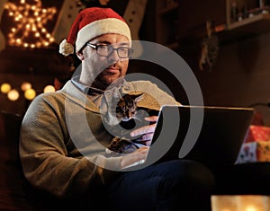 A man in Santa`s New Year hat holds a cat and working with laptop.