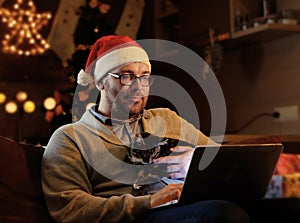 A man in Santa`s New Year hat holds a cat and working with laptop.