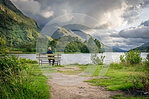 A man sat on bench enjoying a view of a lake and mountains