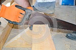 A man sawing a wooden board with a saw, his hands in orange protective gloves, saw in motion; the assistant holds the