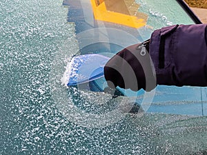 Man scrapes hoarfrost with a plastic scraper from the side window of a blue car. Hand in a black leather glove in the cold.