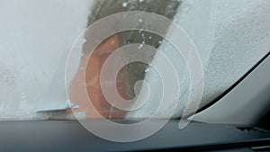 A man scratches the front window of his car on a cold winter morning