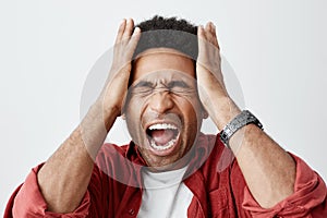 Man screaming from migraine. Close up portrait of black-sinned unhappy guy with afro hairstyle in white t shirt under