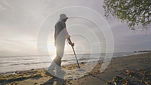 Man searching for valuables under the sand using a metal detector walking with his dog on tropical beach at sunrise.