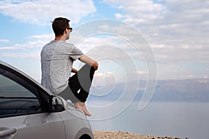 Man seated on the engine hood of a rented car on a road trip in israel