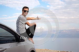 Man seated on the engine hood of a rented car on a road trip in israel