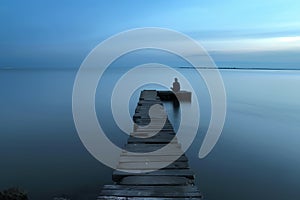 man seated on a jetty extending into calm water at dusk