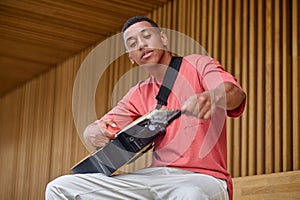 Man seated tuning guitar looking at strings outdoors