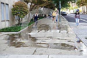 A man is seen power washing a city sidewalk with two girls walking around the area