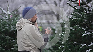 Man selects Christmas tree at Christmastime market and takes tips on correct choice by phone from his family in snowy