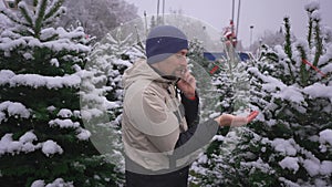 Man selects Christmas tree at Christmastime market and takes tips on correct choice by phone from his family in snowy