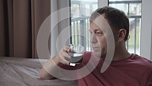 Man with a serious expression is drinking from a glass sitting on the sofa window background